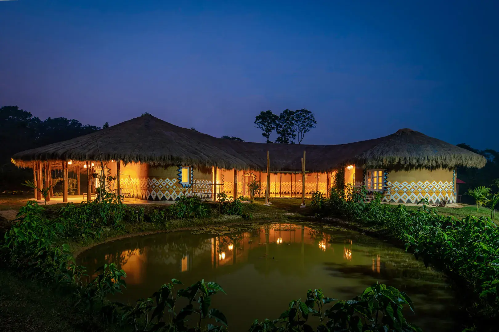 Eco Vacation House at dusk — thatched pavilions glowing across the pond
